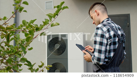 A technician uses a tablet to adjust settings on an external heat pump unit outside a home. A technician uses a tablet to adjust settings on an external heat pump unit outside a home. 118654909