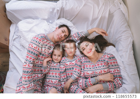 Top view happy family in festive pajamas playing on bed on Christmas Eve in their apartment. Dad, mom,daughter, son are lying on bed and having fun together. Family celebrating winter holidays at home 118655573