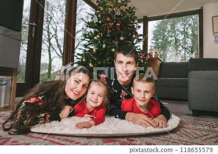 Smiling family portrait in festive sweaters laying under decorated Christmas tree at home during holiday. Happy parents and kids cuddling, feeling excited. Family time together. Happy childhood. Smiling family portrait in festive sweaters laying under decorated Christmas tree at home during holiday. Happy parents and kids cuddling, feeling excited. Family time together. Happy childhood. 118655579