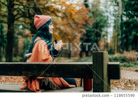 Smiling Woman wrapped in Shawl Poncho enjoying hot drink from thermos cup sitting on the bench during autumn walk in fall forest. Feeling harmony, relax, personal fulfilment. Local travel lifestyle. 118655590