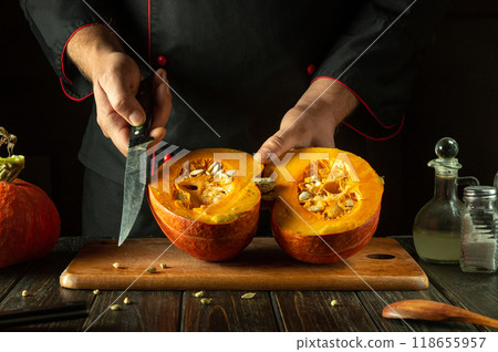 Chef cuts a large pumpkin into two halves before cooking it on the kitchen table. Concept of cooking vegetable dish with pumpkin and spices Chef cuts a large pumpkin into two halves before cooking it on the kitchen table. Concept of cooking vegetable dish with pumpkin and spices 118655957