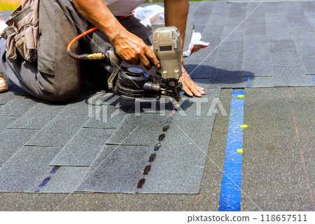 Construction worker used an air pneumatic nail gun when installing new bitumen shingles on roof Construction worker used an air pneumatic nail gun when installing new bitumen shingles on roof 118657511