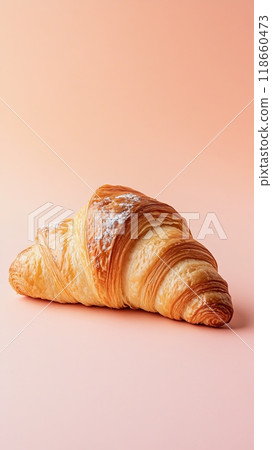 A golden, flaky croissant is placed against a soft pastel peach background 118660473