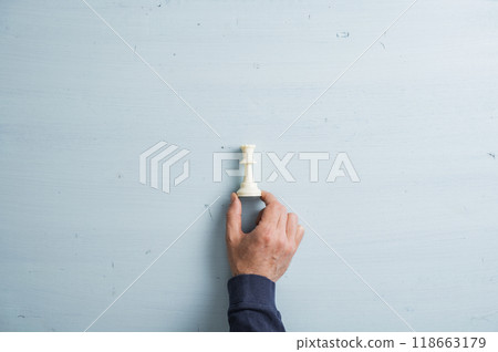 Male hand placing white king chess figure on a plain simple light blue wooden background Male hand placing white king chess figure on a plain simple light blue wooden background 118663179