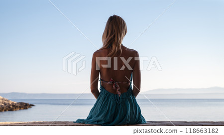 Young woman in blue summer dress sitting by the sea looking into the distance Young woman in blue summer dress sitting by the sea looking into the distance 118663182
