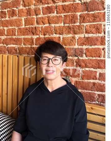 Confident middle-aged woman with glasses sitting against a coffeeshop brick wall. Mature female in casual black hoodie enjoying a relaxing moment Indoors. Confident middle-aged woman with glasses sitting against a coffeeshop brick wall. Mature female in casual black hoodie enjoying a relaxing moment Indoors. 118663280