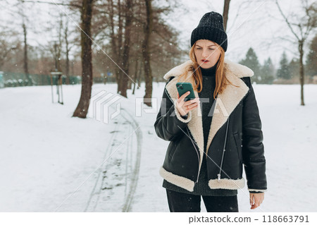 Annoyed upset blonde woman looking at her smart phone with frustration outdoors. Beautiful woman walking in the park on a winter snowy day 118663791