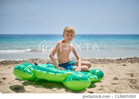 Idyllic Family Getaway: Joyful Boy Lounging on Whimsical Turtle-Shaped Float Amidst Golden Sands and Tranquil Ocean Waves 118665008