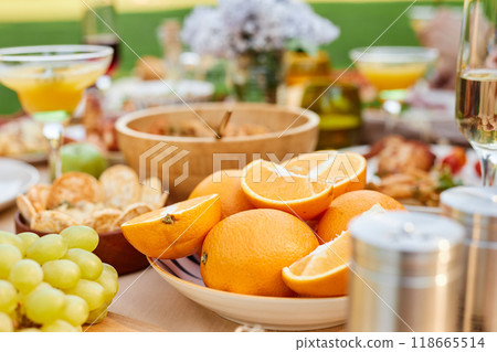 Fresh oranges arranged in a white bowl on a wooden table surrounded by a variety of foods and drinks, creating a vibrant and festive outdoor gathering setup Fresh oranges arranged in a white bowl on a wooden table surrounded by a variety of foods and drinks, creating a vibrant and festive outdoor gathering setup 118665514