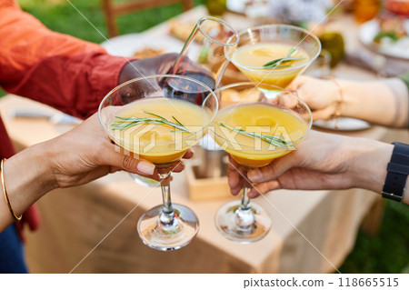 Hands raised clinking cocktail glasses garnished with rosemary twigs while gathering at outdoor celebration event with blurred background 118665515