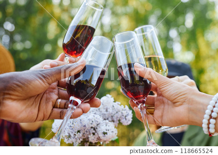 Close-up of hands raising wine glasses in toast during outdoor celebration with flowers in background creating festive atmosphere Close-up of hands raising wine glasses in toast during outdoor celebration with flowers in background creating festive atmosphere 118665524
