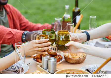 Group of friends toasting glasses at outdoor picnic table with food and drinks, celebrating togetherness and enjoying delicious meal outside on sunny day 118665530