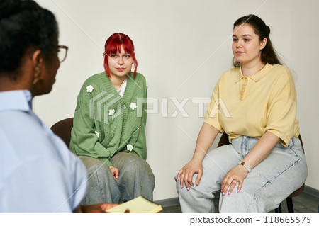 Three individuals engaging in therapy session, including therapist and two women on chairs. One woman has red hair while the other has brown hair, both wearing casual clothes Three individuals engaging in therapy session, including therapist and two women on chairs. One woman has red hair while the other has brown hair, both wearing casual clothes 118665575