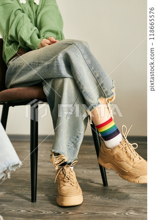 Person seated on chair wearing rainbow socks and beige sneakers with crossed legs, casual denim jeans frayed at the hem and green sweater, relaxed and casual atmosphere Person seated on chair wearing rainbow socks and beige sneakers with crossed legs, casual denim jeans frayed at the hem and green sweater, relaxed and casual atmosphere 118665576