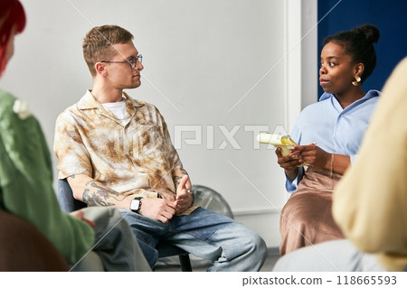 Two colleagues engaging in conversation about project details in comfortable office setting. One holding notebook while other listens attentively wearing casual attire Two colleagues engaging in conversation about project details in comfortable office setting. One holding notebook while other listens attentively wearing casual attire 118665593