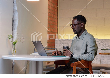 Man wearing light gray shirt sitting at white desk working on laptop and holding reusable cup of coffee with empty brick wall in background 118665656