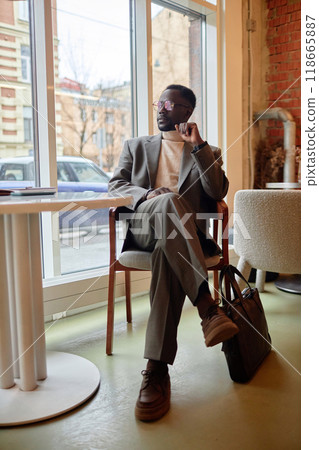 African American man wearing stylish beige suit sitting comfortably in modern cafe with large windows. Showcasing a sophisticated and relaxed demeanor while gazing thoughtfully outside 118665887