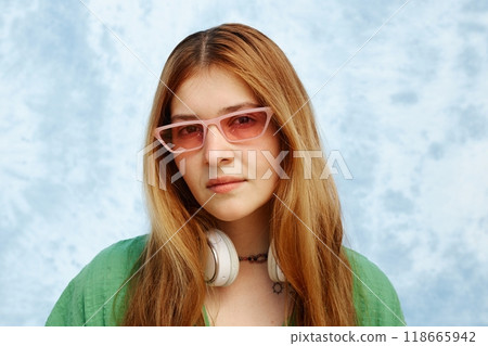 Portrait of young Hispanic woman with long hair wearing pink glasses and white headphones, standing against blue textured background Portrait of young Hispanic woman with long hair wearing pink glasses and white headphones, standing against blue textured background 118665942