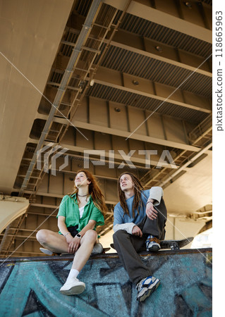 Two friends sitting under bridge enjoying sunny day while relaxing on ledge. Both individuals are looking up at sky, seemingly lost in thought Two friends sitting under bridge enjoying sunny day while relaxing on ledge. Both individuals are looking up at sky, seemingly lost in thought 118665963