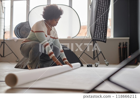 Side view portrait of young Black woman working as photographer and unrolling paper background on floor preparing for studio photoshoot copy space Side view portrait of young Black woman working as photographer and unrolling paper background on floor preparing for studio photoshoot copy space 118666211