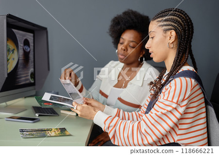 Side view portrait of two creative African American women discussing photos in studio and holding art prints for selection Side view portrait of two creative African American women discussing photos in studio and holding art prints for selection 118666221