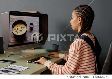 Side view portrait of young Black woman with cornrows hairstyle as female photographer using computer and editing food photography images on screen 118666230