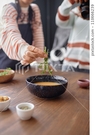 Vertical closeup of women working on food photography photoshoot in studio with focus on hand putting green decor on plate of cream soup copy space 118666239