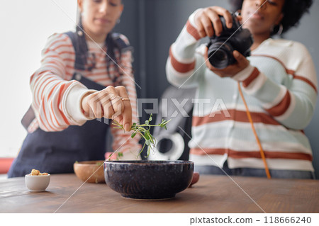 Two Black young women working on food photography photoshoot together in studio with focus on hand putting green decor on plate copy space Two Black young women working on food photography photoshoot together in studio with focus on hand putting green decor on plate copy space 118666240