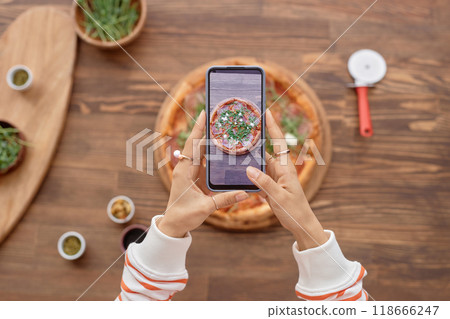 Top view of female hands holding smartphone and taking photo of delicious pizza on wooden table during mobile food photography set copy space 118666247