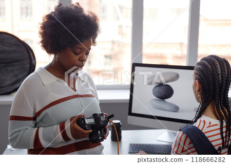 Portrait of young Black woman as female photographer holding camera and looking at photos with colleague during product photography photoshoot in studio 118666282