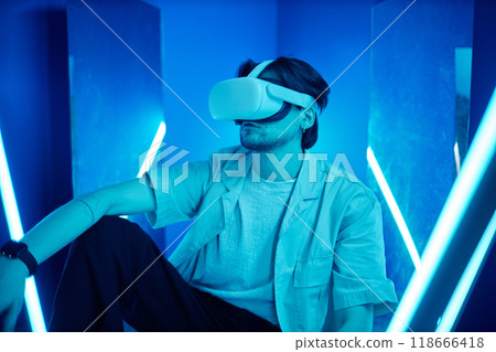Portrait of man wearing VR headset sitting on floor in neon lights experiencing digital cyberspace 118666418