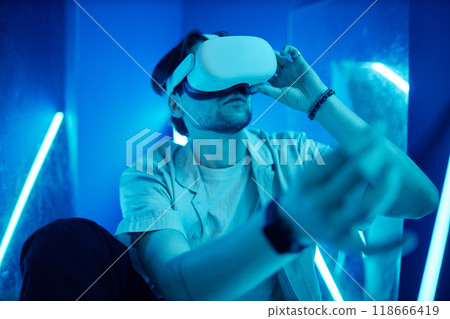 Portrait of man wearing VR headset sitting on floor in blue neon lights and reaching out experiencing digital cyberspace 118666419
