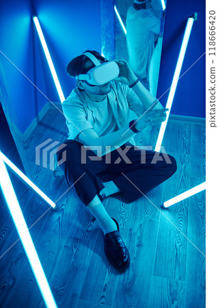 Vertical full length portrait of man wearing VR headset sitting on floor in blue neon lights and reaching out 118666420