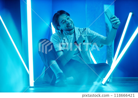 Full length portrait of smiling man doing livestream with smartphone while sitting on floor in room with blue neon lights 118666421