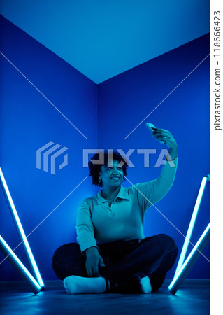 Full length portrait of African American woman taking selfie photo with smartphone while sitting on floor in room with blue neon lights 118666423