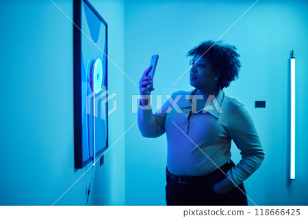 Side view portrait of African American woman looking at paintings in blue neon light during modern art show and taking pictures, copy space 118666425