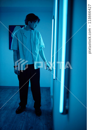 Vertical side view portrait of young man interacting with blue neon light strips during modern art show in museum enjoying multisensory experience 118666427