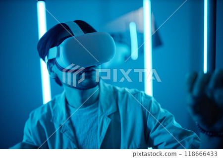 Portrait of young man wearing VR headset in modern art show interacting with blue neon lights enjoying digital mixed reality experience 118666433