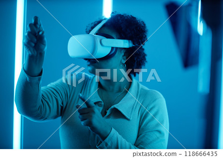 Side view portrait of Black woman wearing VR headset in modern art show interacting with blue neon lights experiencing digital cyberspace Side view portrait of Black woman wearing VR headset in modern art show interacting with blue neon lights experiencing digital cyberspace 118666435