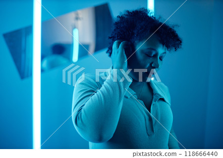Minimal waist up portrait of African American woman in room with blue neon lights listening to in ear music, modern art show and digital cyberspace concept 118666440