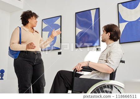 Side view portrait of smiling Black woman talking to man using wheelchair in accessible modern art gallery and discussing abstract paintings 118666511