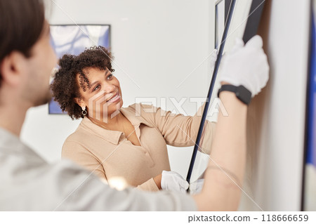 Side view portrait of smiling Black woman hanging painting on wall in modern art gallery or museum with colleague 118666659