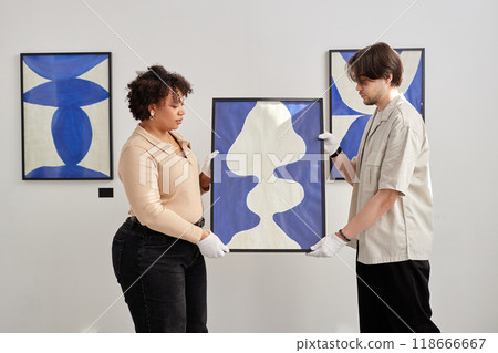 Side view of two people holding abstract painting with blue shapes while preparing for exhibition in modern art gallery Side view of two people holding abstract painting with blue shapes while preparing for exhibition in modern art gallery 118666667