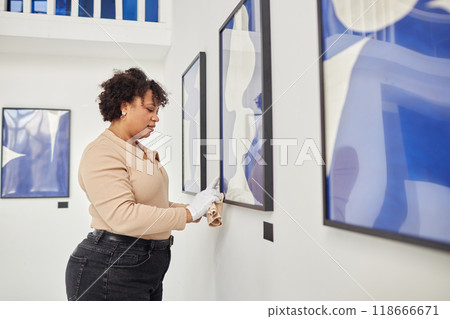 Side view portrait of Black adult woman caring for abstract paintings in modern art gallery and wearing white gloves copy space 118666671
