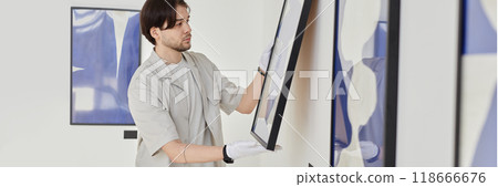 Header image of young man hanging abstract art pictures on wall while planning modern art gallery exhibition copy space Header image of young man hanging abstract art pictures on wall while planning modern art gallery exhibition copy space 118666676