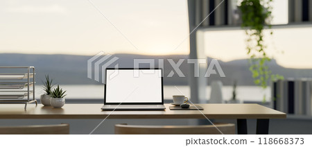 A laptop on a wooden desk in a modern office, featuring a large window with a nature view. 118668373