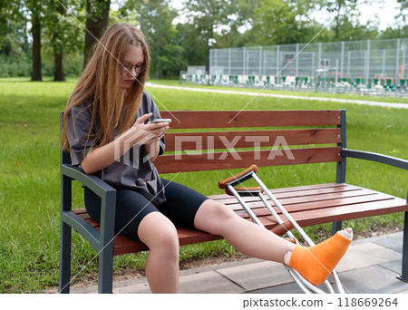 Teenage girl with a leg cast sitting on a park bench, using her smartphone to engage with social media as she recovers in the summer setting Teenage girl with a leg cast sitting on a park bench, using her smartphone to engage with social media as she recovers in the summer setting 118669264