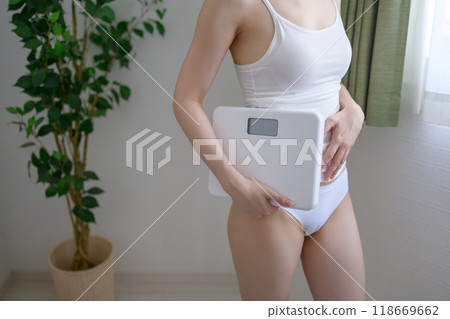 一位穿著內衣的年輕苗條酷女人站在窗邊，手裡拿著體重秤，體重管理，明亮的自然光。 118669662