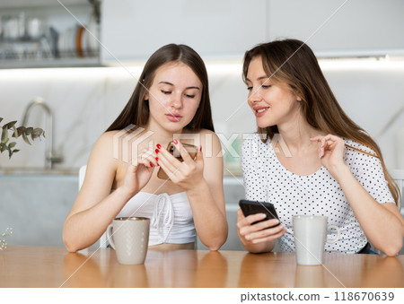 Happy mother and teen daughter looking at their phones sitting at kitchen table Happy mother and teen daughter looking at their phones sitting at kitchen table 118670639