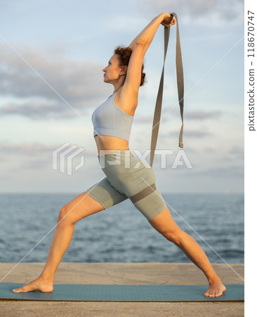 Yoga practice on embankment, female athlete fulfil carry out warrior pose with belt Yoga practice on embankment, female athlete fulfil carry out warrior pose with belt 118670747